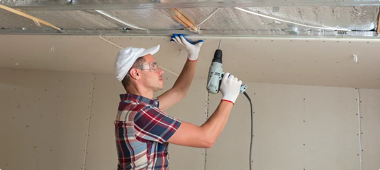 Worker screwing drywall to ceiling with power drill during construction
